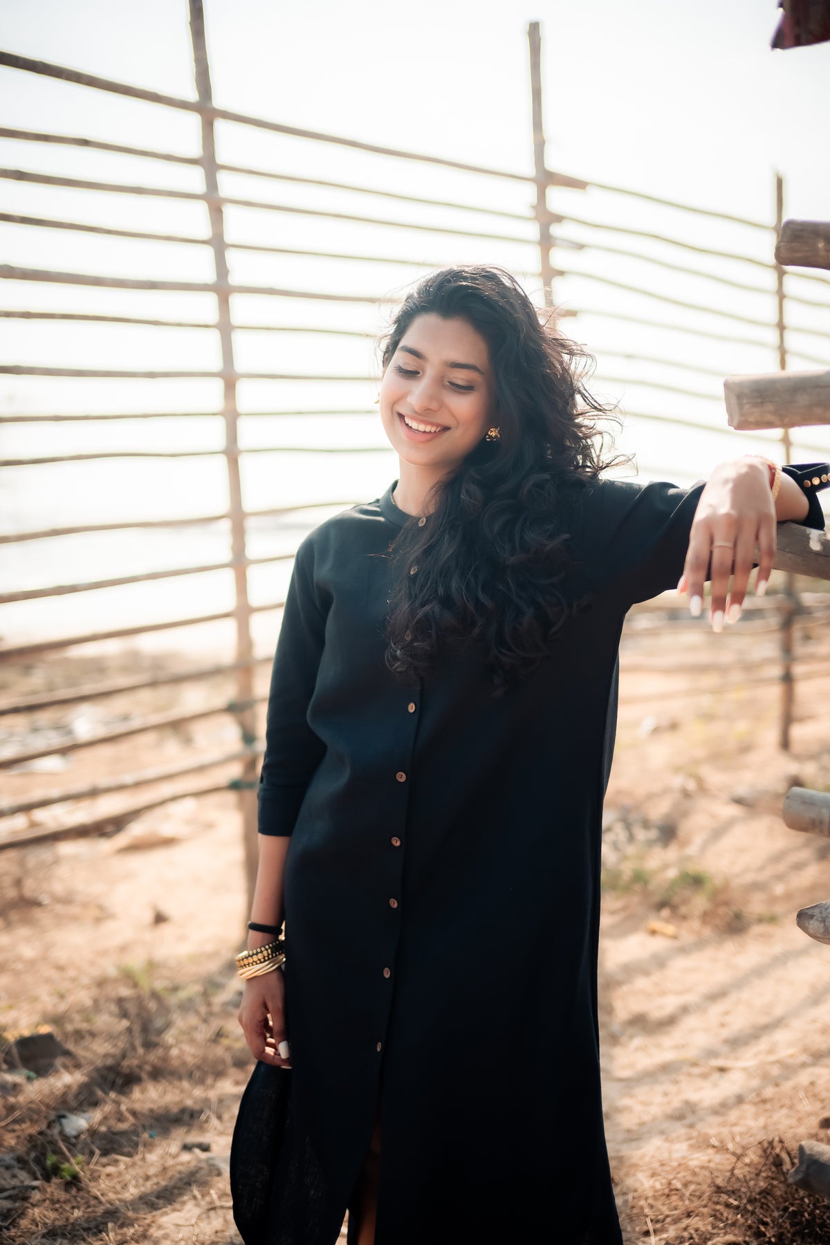 A South Asian woman smiling softly with her eyes looking down, wearing a long black button-down kurta or tunic dress with gold bangles on her wrist. She leans lightly against a rustic wooden fence in a sunny, sandy outdoor environment.