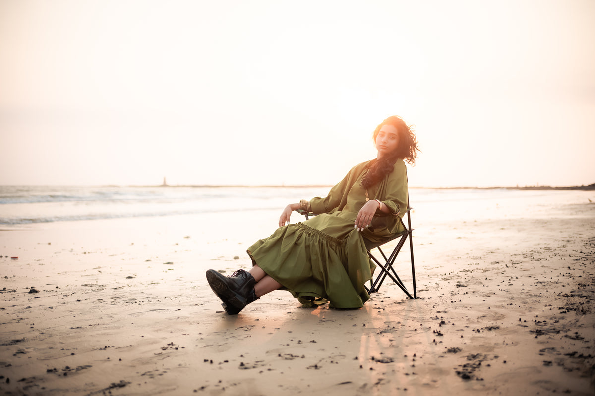 A South Asian woman relaxing on a folding camping chair on a sandy beach during sunset, wearing a long olive green dress paired with black combat boots. The golden sunlight casts a soft glow over the ocean waves in the background.