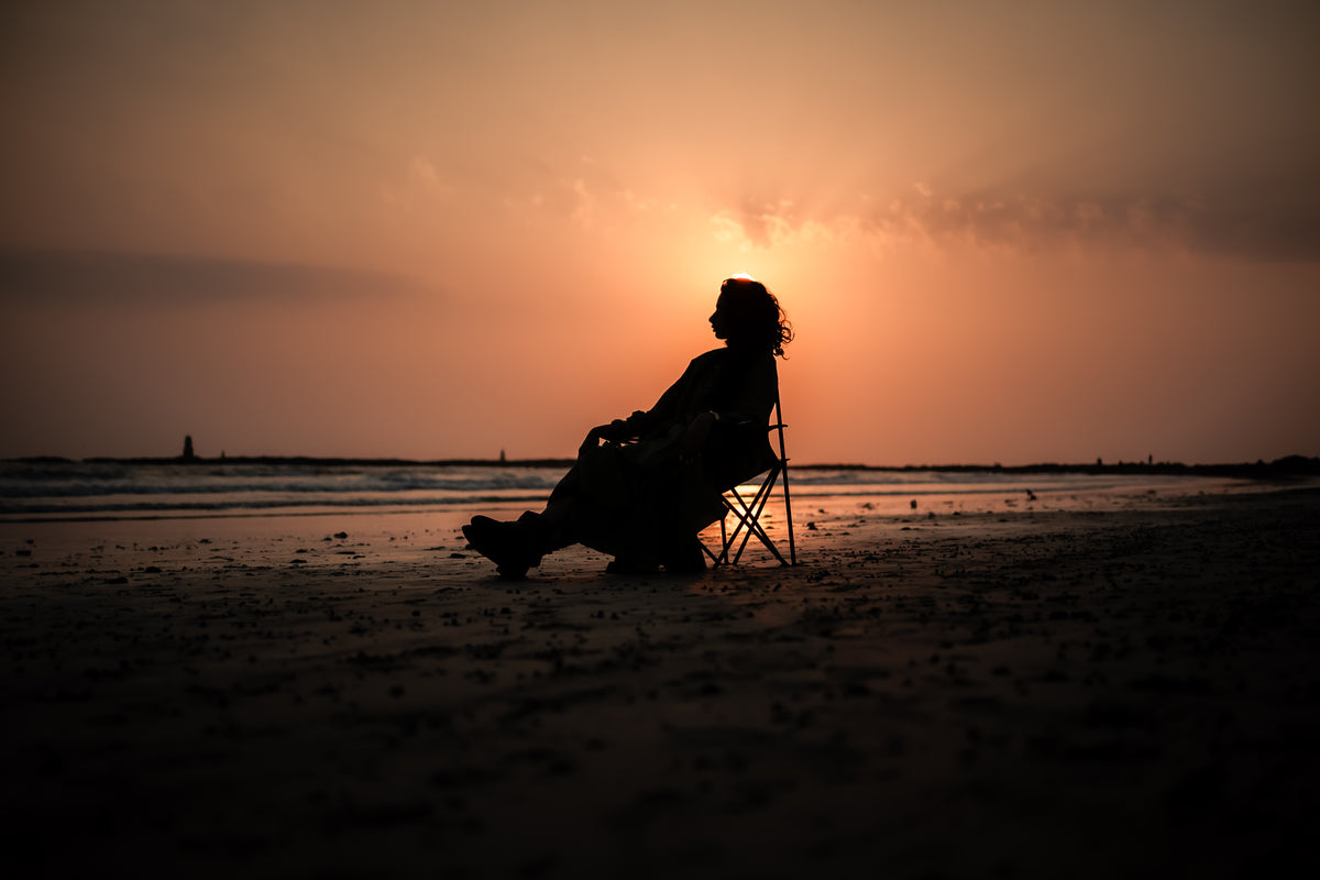 A dark silhouette of a young woman sitting in a folding camping chair on a sandy beach during sunset, with the glowing orange sun positioned directly behind their head, creating a radiant halo effect over the calm ocean waves.